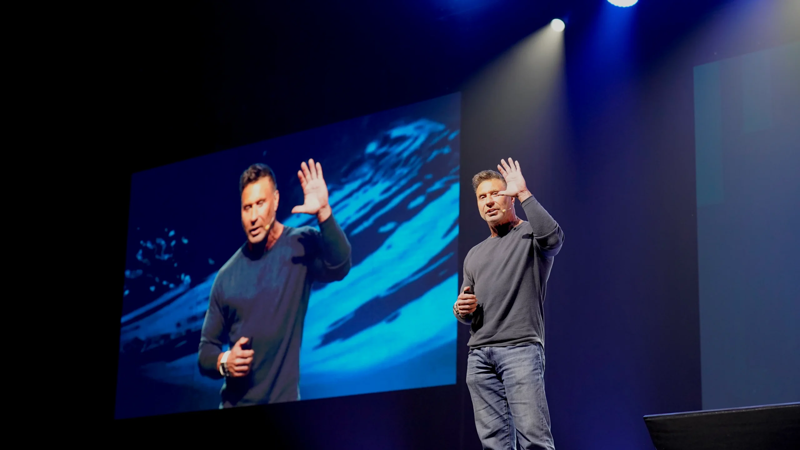 Life Surge Speaker Ed Mylett stands on the stage with his image on a screen behind him